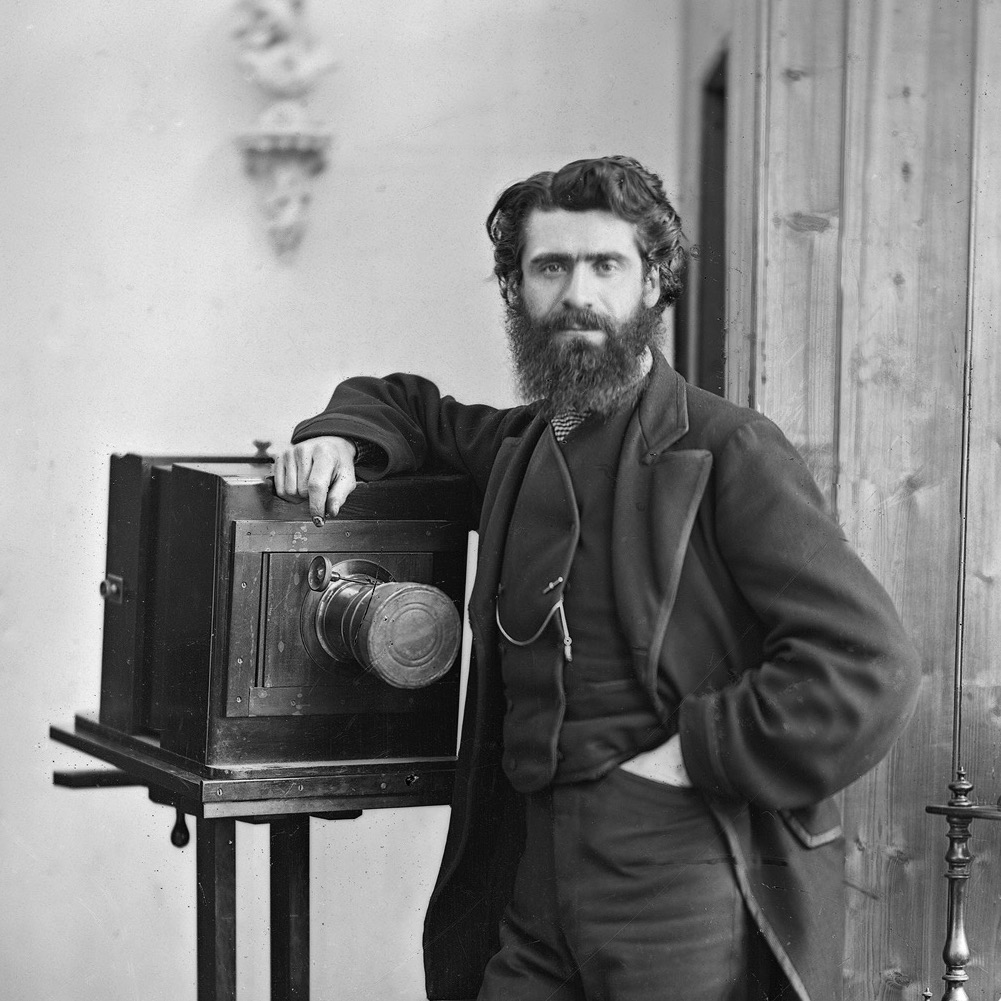 A bearded man in jacket and waistcoat standing by a large camera. 